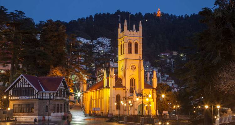 Illuminated yellow Christ Church on Shimla Mall Road with hillside houses behind at dusk.