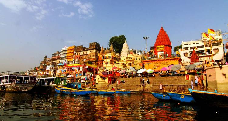 Colorful ghats and temples of Varanasi lined along the Ganges River with boats in foreground.