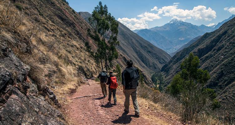 Hikers follow a red-dirt trail through a dramatic Andean valley with snow-capped peaks ahead.