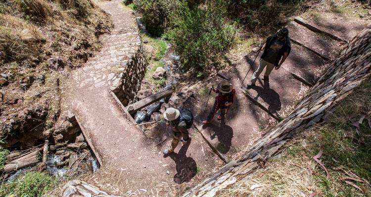 Overhead view of trekkers crossing a small stream on a rustic stone and log bridge within a canyon.