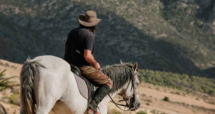 Einsamer Reiter mit Cowboyhut auf einem grauen Pferd betrachtet zerklüftete Mittelmeerhügel.