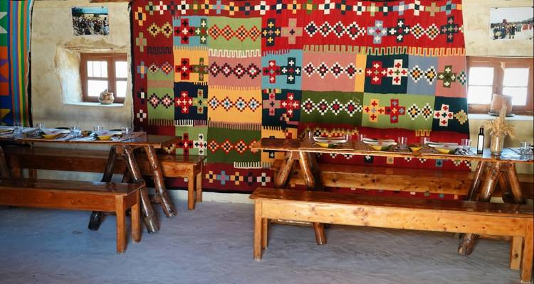 Bunte gewebte Wandteppiche schmücken eine schlichte Speisehalle mit langen Holzbänken und Tischen.