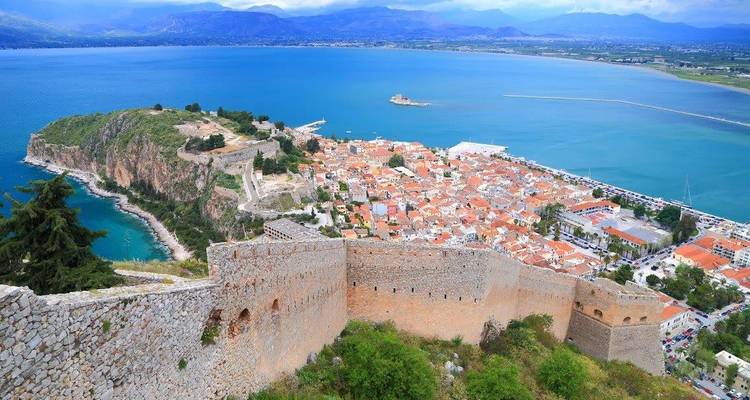 A high-angle view of Nafplion showing the coastal fortress walls, terracotta-roofed old town and turquoise sea backed by distant mountains.