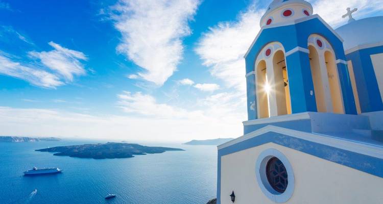 Sunlit blue-and-cream bell tower of a Santorini church with caldera and cruise ships below.