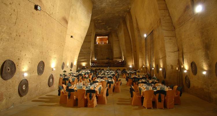 Large subterranean hall set for a banquet with round tables covered in white and ochre linens.