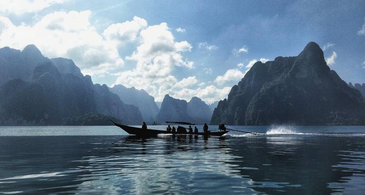 Long-tail boat carrying passengers across a serene lake framed by dramatic limestone karsts under bright clouds in Khao Sok.