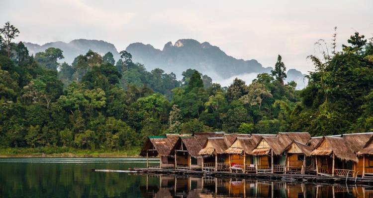 Rows of thatched floating bungalows rest on a calm emerald lake with jungle-covered mountains rising behind them.