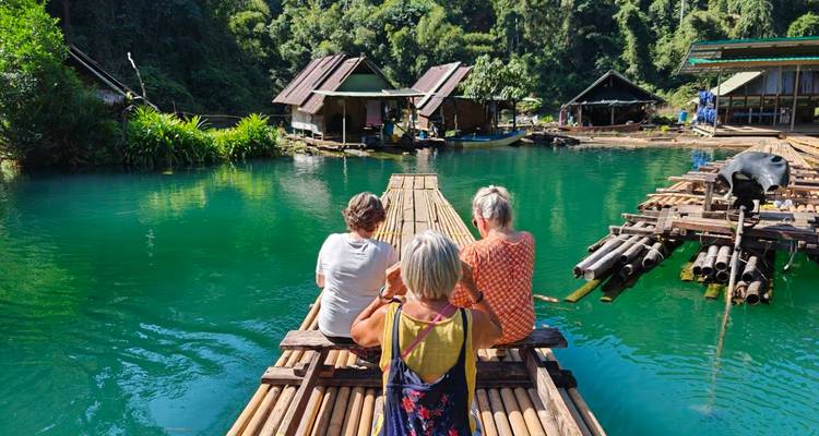 Three travellers sit on a bamboo raft, gazing over an emerald lagoon and floating houses.