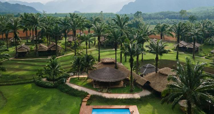Vista aérea de un eco-resort rodeado de palmeras con una piscina central ubicada entre campos verdes ondulantes y colinas distantes.