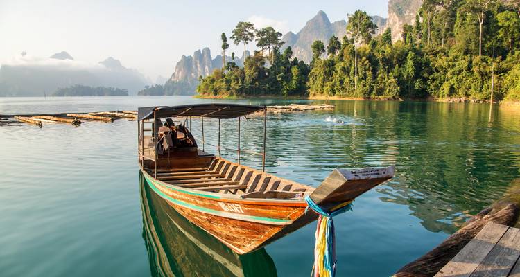 Un bote tradicional de cola larga flota en un lago quieto como un espejo rodeado de picos calizos brumosos al amanecer.