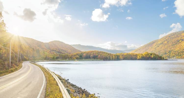 Une route côtière ensoleillée serpente le long d'un lac encadré par un feuillage automnal dans les Hautes-Terres du Cap-Breton.