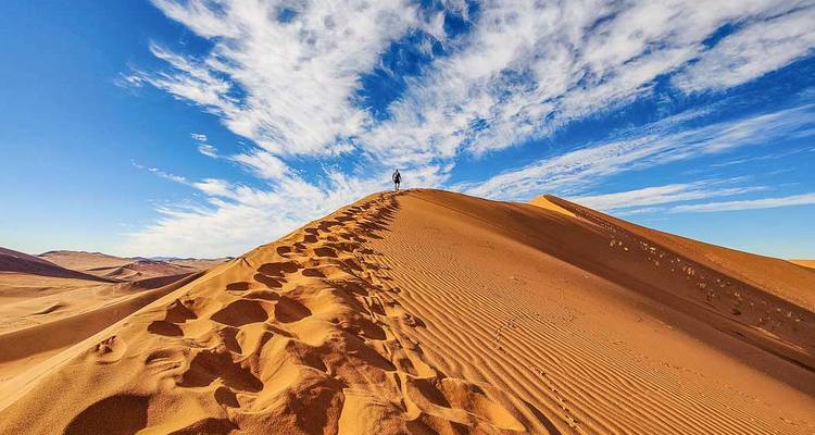 Un voyageur solitaire marche le long de la crête d'une haute dune de sable orange sous des traînées de nuages spectaculaires.