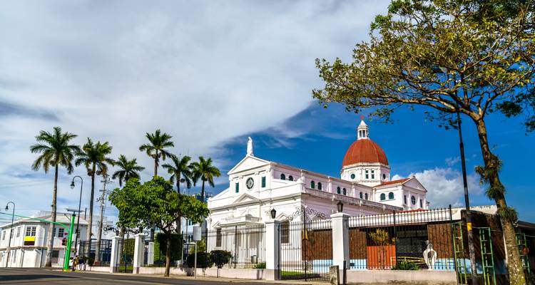 Cathédrale néoclassique blanche avec dôme rouge se détachant sur un ciel bleu vif du Costa Rica et des palmiers.
