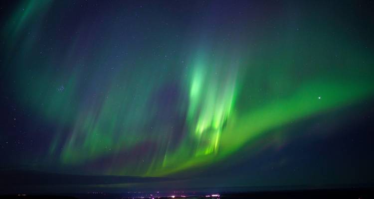 Vivid green aurora borealis dances across a starry night sky above distant town lights