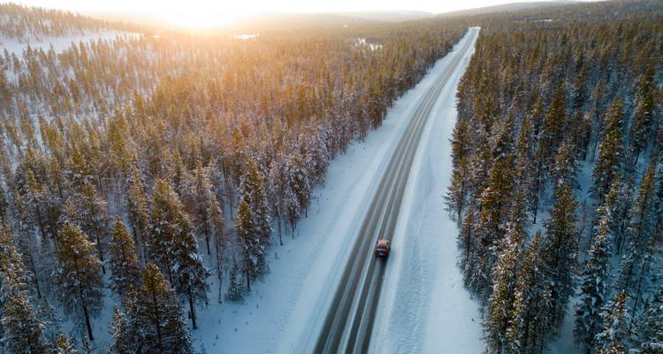 Aerial winter road cutting through dense snow-covered forest with sunrise glow on the horizon