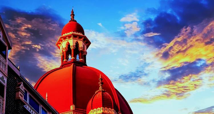 Red domes of the historic Taj Mahal Palace hotel glowing at sunset against dramatic clouds.