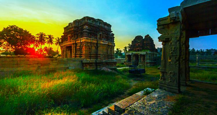 Ancient stone temples of Hampi bathed in warm sunrise light with palm trees beyond.
