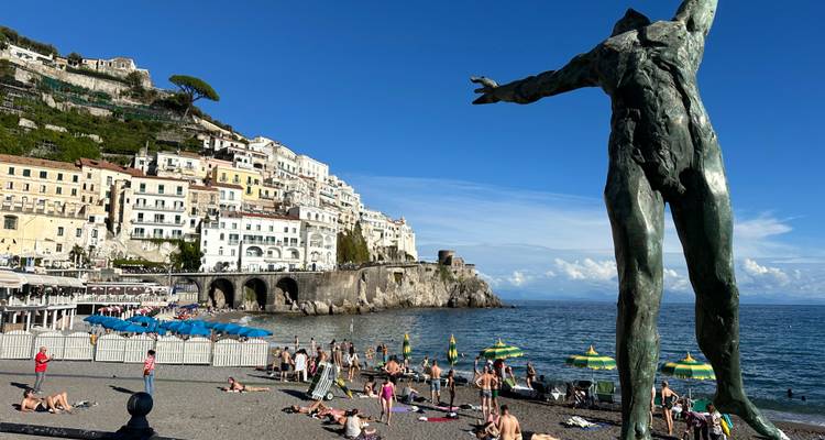Beach scene at Amalfi with sunbathers, a massive bronze statue in foreground and cliffside town behind.