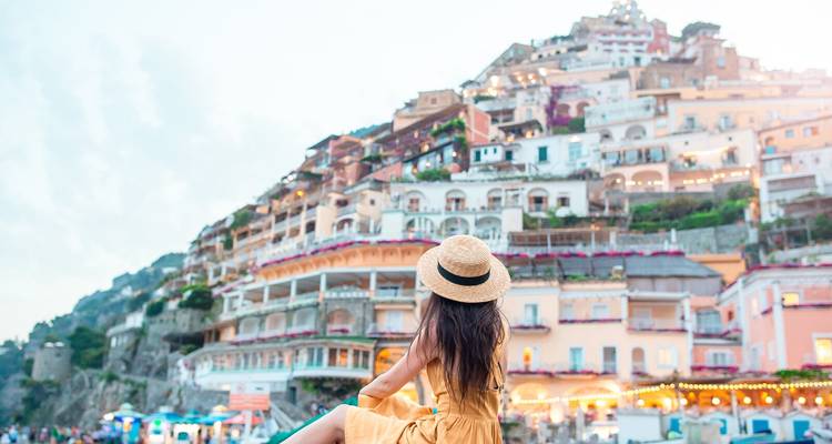 Woman in a straw hat and yellow dress gazing at the pastel houses of Positano from the beach at dusk.
