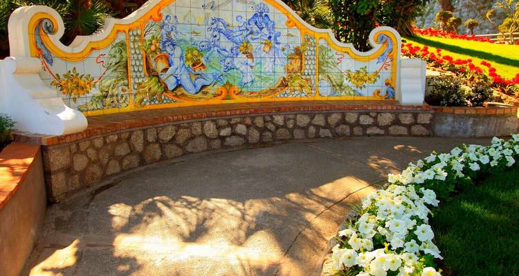 Colorful ceramic-tiled bench set in a sunny Mediterranean garden with bright flowers and ornate stonework.