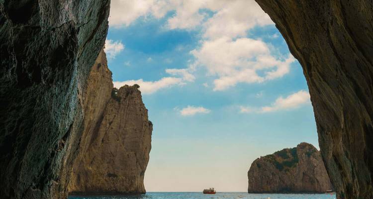 Sea cave framing a view of the Faraglioni rock stacks rising from the blue waters under a bright sky.