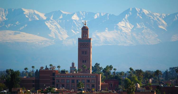 Koutoubia Mosque rising over Marrakesh rooftops with snow-capped Atlas Mountains in the background.