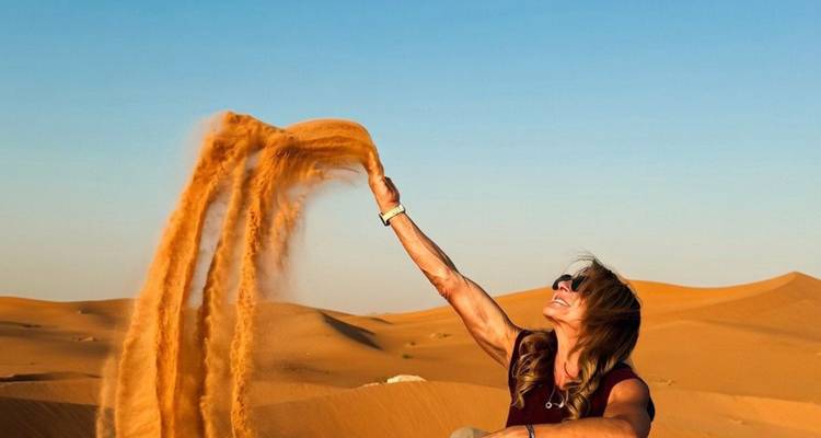 Smiling woman tossing a cascade of golden sand against a clear desert sky.