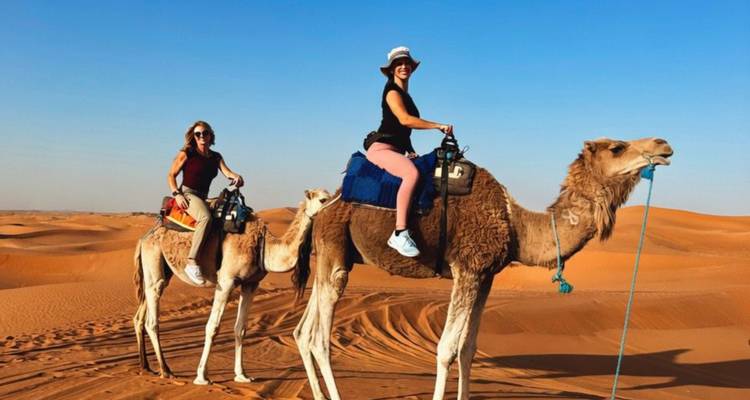 Two women ride camels across rolling orange dunes under a bright blue sky.