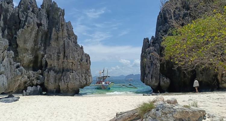 Étroite brèche sablonneuse entre des rochers déchiquetés s'ouvrant sur des eaux turquoise avec un bateau au-delà