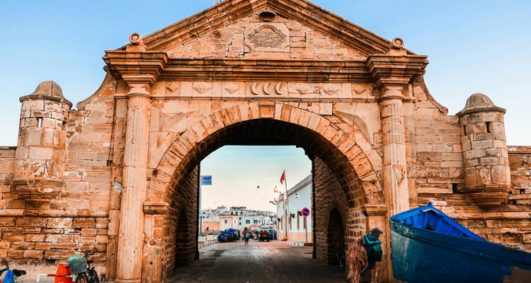 Stone arch gateway of Essaouira's seaside fort with a blue boat nearby at golden hour.
