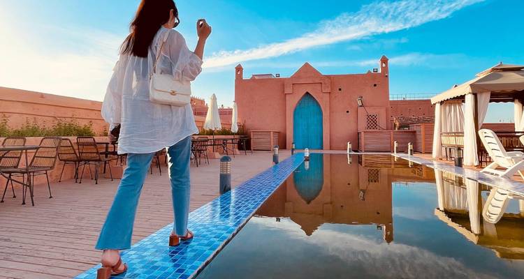 Une jeune femme en vêtements clairs passe devant un bassin réfléchissant sur une terrasse de toit avec des murs marocains roses et une porte bleu vif.