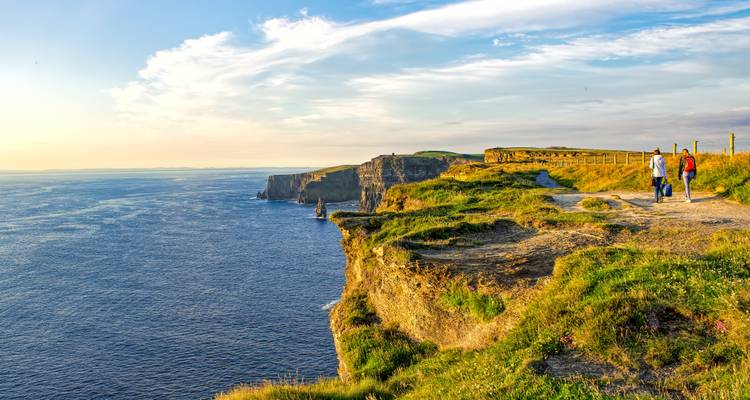 Hikers walking along grassy cliff edge overlooking vast Atlantic Ocean and famous sea stacks.
