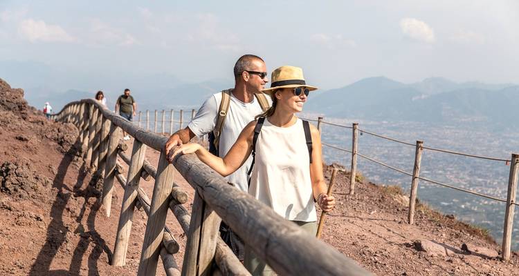 Couple hiking along the wooden railing on the rim of Mount Vesuvius with hazy mountains in the background.