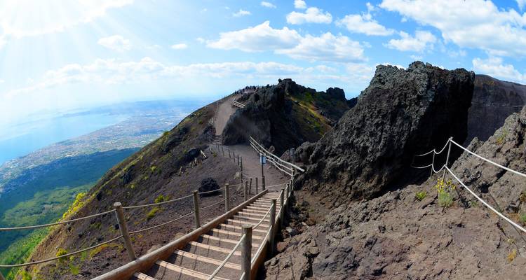 Wooden stairs lead along the rocky rim of a volcanic crater with sweeping views over the distant coastline and blue sea.