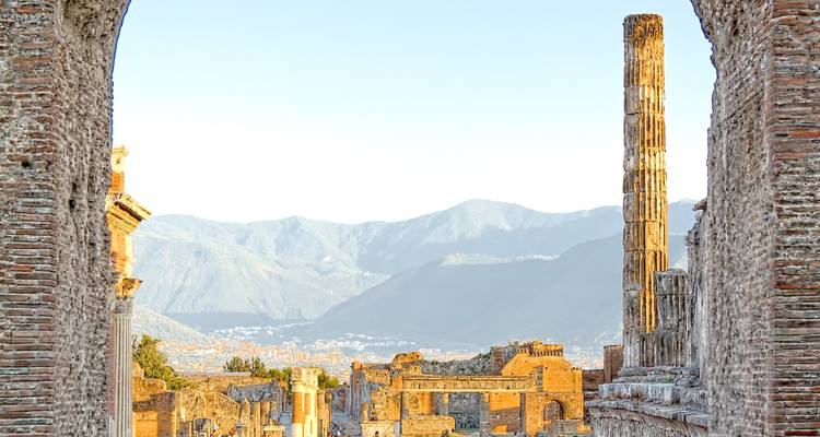 Sunlit stone columns and ruins of ancient Pompeii framed by brick archway with mountains behind.
