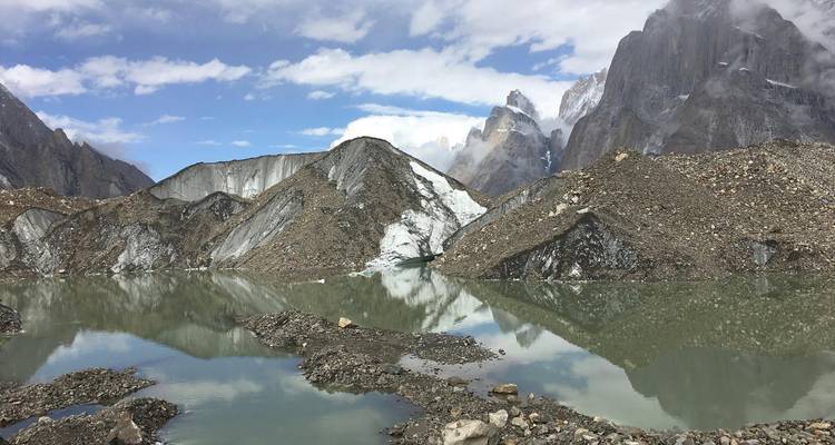 Melkachtig gletsjermeer dat met puin bedekte ijswanden en grillige granietrotsen weerkaatst in bewolkt licht.