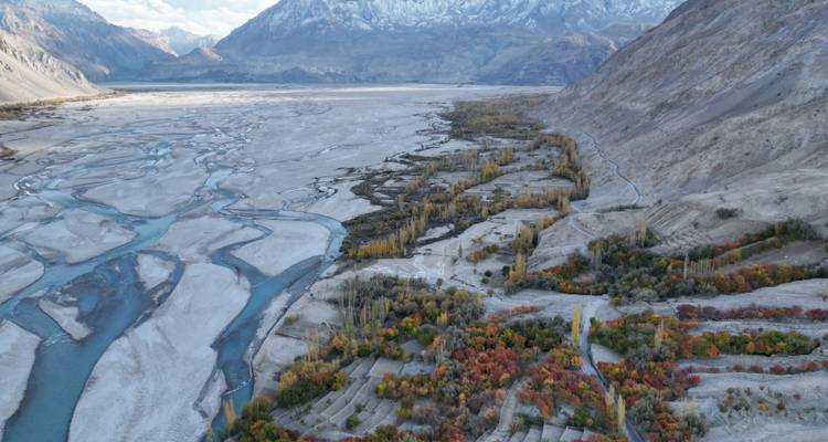 Luchtfoto van een brede gevlochten riviervallei met bomen in herfstkleuren tegen ruige bergen in de verte.