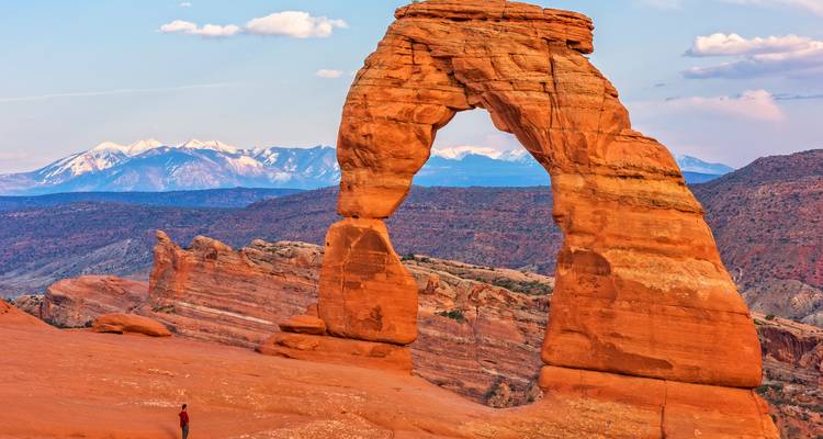 A lone traveler gazes up at Delicate Arch with snow-capped mountains in the distance under pastel evening light.