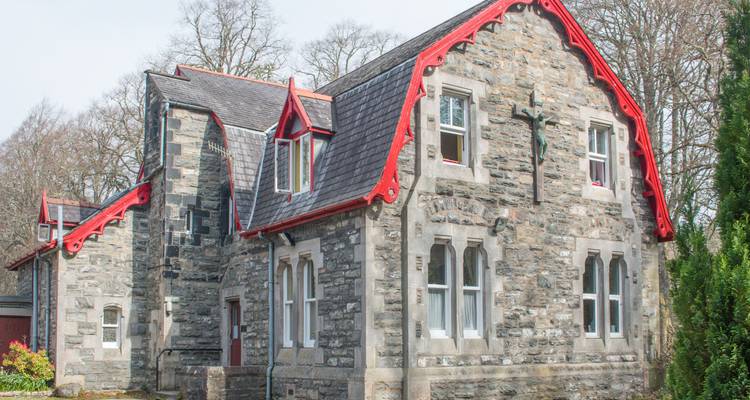 Stone cottage with red trim and a cross on the façade set against bare winter trees