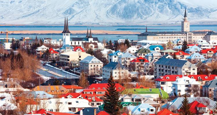 Colourful low-rise city with pointed church spires set before snow-covered mountains and a broad bay.