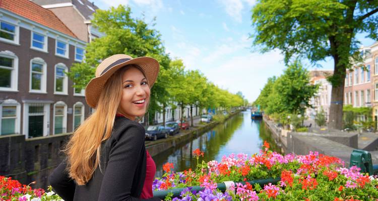 Glimlachende reiziger met zonnehoed staat op een met bloemen versierde brug over een Nederlandse gracht.