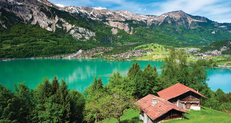 Un lac alpin d'un turquoise intense reflète les versants boisés et les pics enneigés, avec des cabanes rustiques parsemant une pente herbeuse.