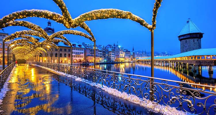 Evening view of Lucerne’s illuminated Chapel Bridge and Water Tower over calm river with snowy edges.