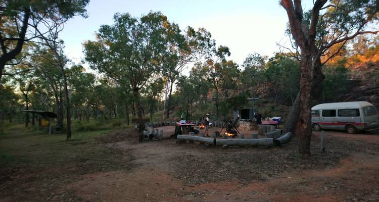 Bush campsite with a fire ring, picnic tables and a lone camper preparing dinner.