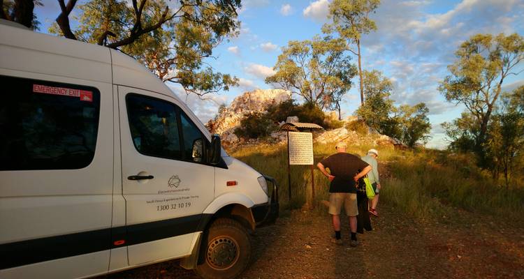 Tour van parked beside an informational sign as two travellers read before a rocky hill.