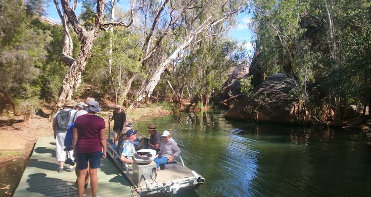 Travellers board a small flat-bottom boat from a wooden jetty on a shaded creek.