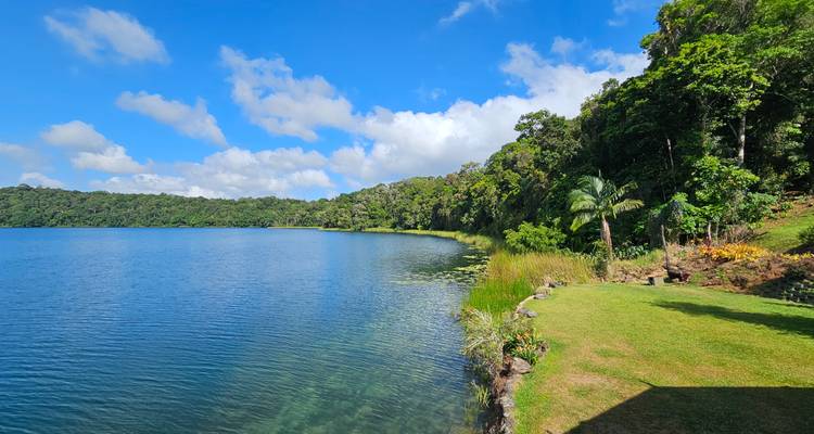 Lac d'un bleu cristallin bordé par une forêt tropicale luxuriante et une pelouse herbeuse sous des nuages gonflés.