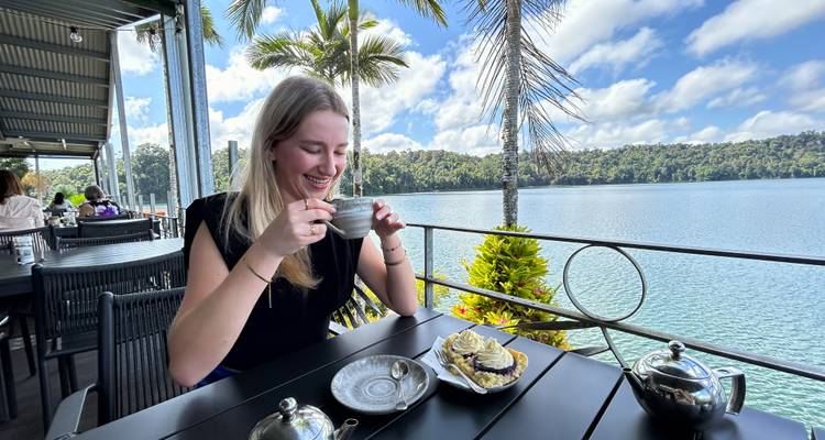 Une femme savoure le thé et le gâteau sur le balcon d'un café au bord du lac, avec des palmiers et une eau bleue calme au-delà.