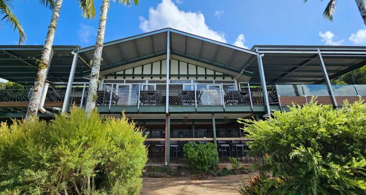Lodge moderne au bord du lac avec de larges vérandas et un toit à pente raide entouré d'arbustes tropicaux sous un ciel bleu.