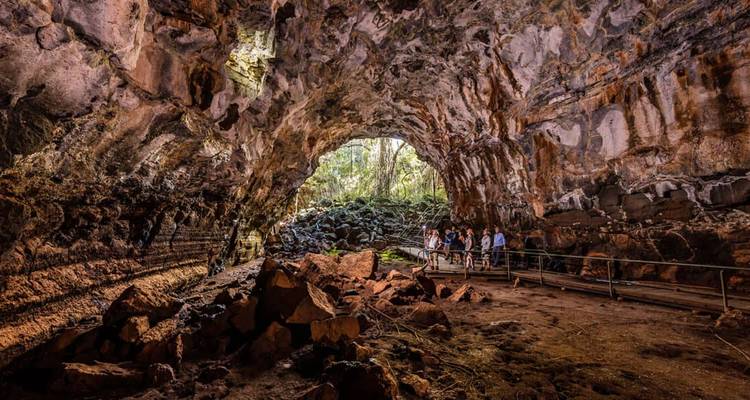 Grande caverne de style tube de lave avec des parois rocheuses escarpées et un groupe de visiteurs se tenant sur une passerelle près de l'ouverture illuminée.